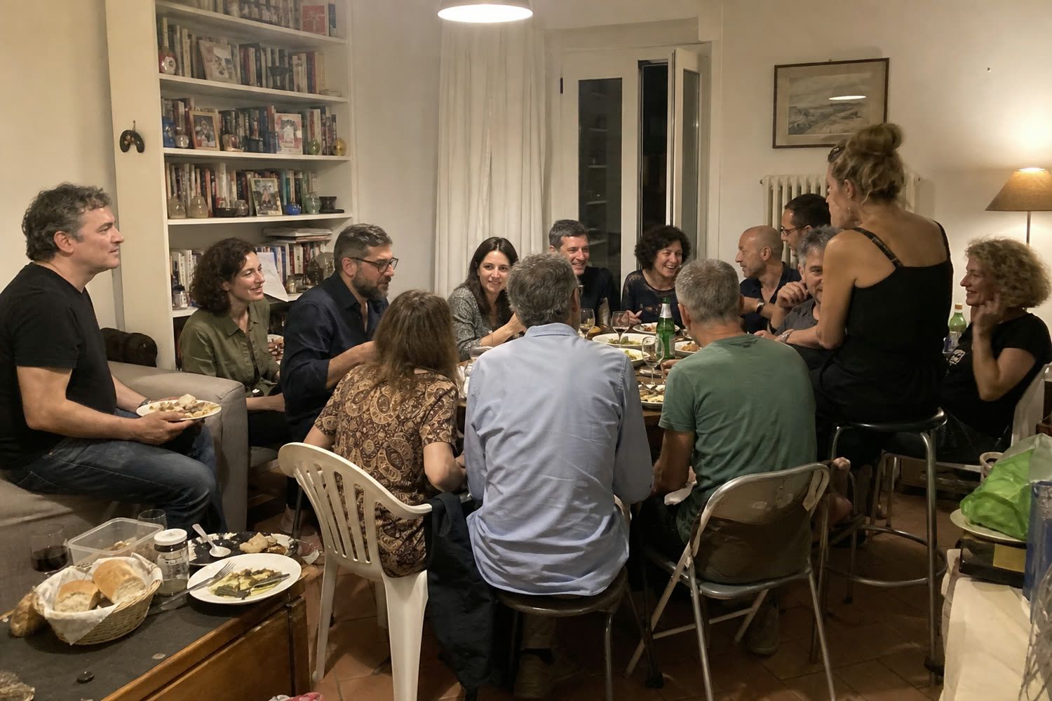 12 persone strette attorno a un tavolo piccolo, tra sedie di plastica, sedie pieghevoli, uno sgabello alto e il bracciolo del divano